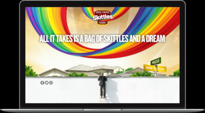 A person on a ladder looks at a rainbow above houses, promoting a Skittles game with the text "All it takes is a bag of Skittles and a dream.