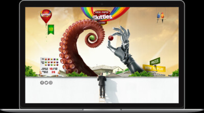 A person climbs a ladder towards a giant octopus tentacle and a robotic hand, promoting a Skittles game.